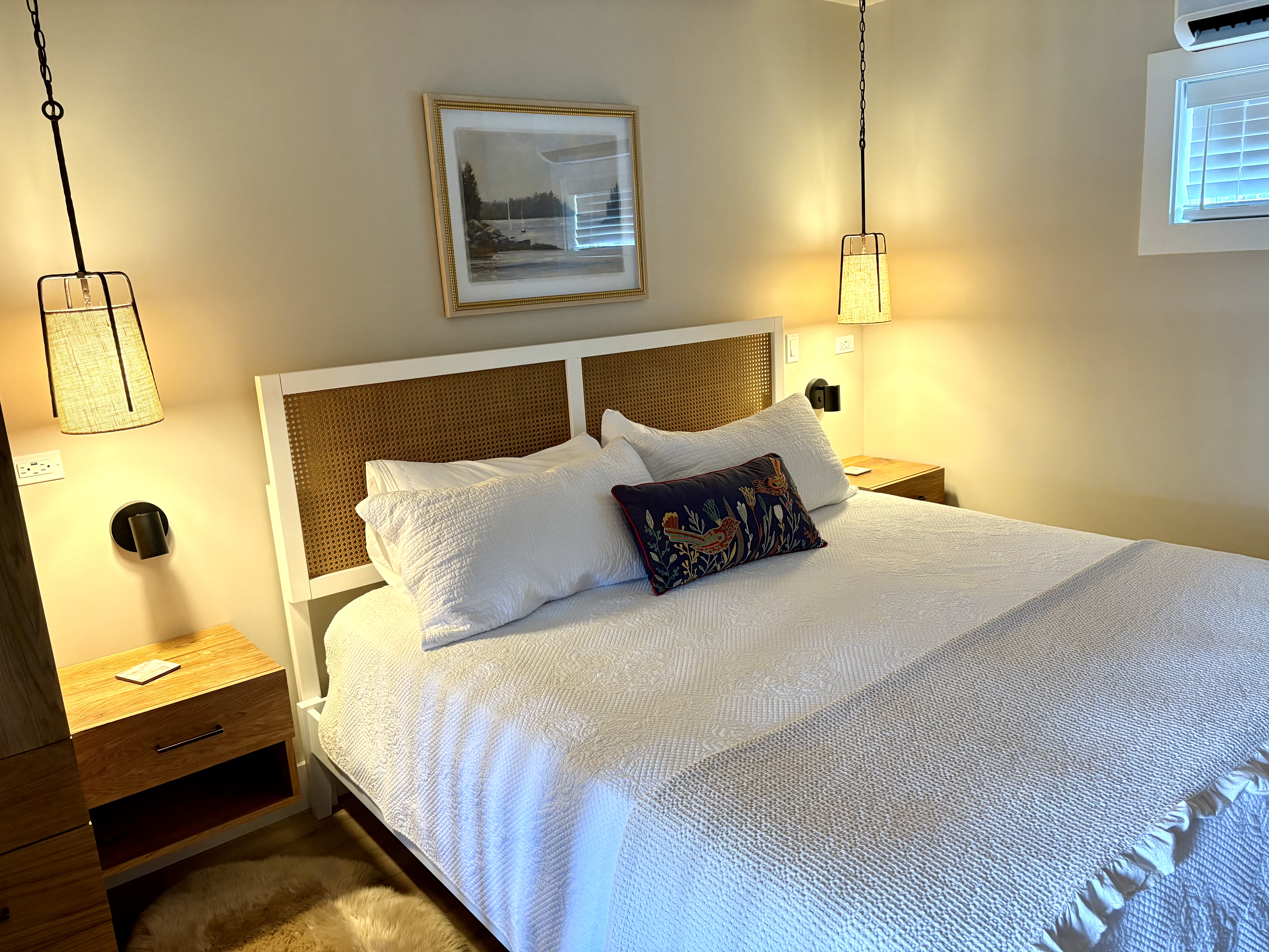 A cozy, well-lit bedroom features a king bed with white linens, a woven headboard, matching wood nightstands, and stylish hanging pendant lights.