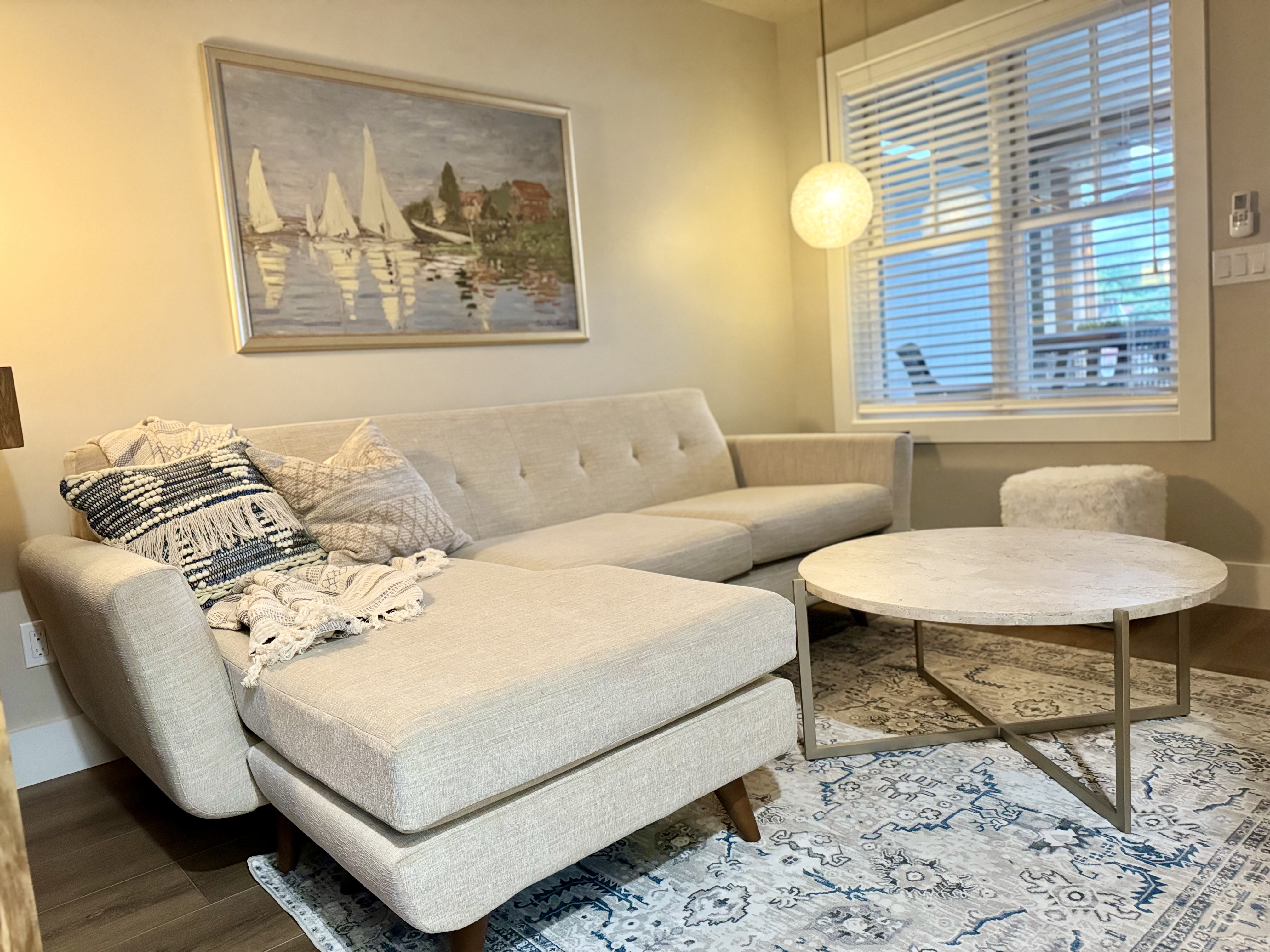A bright living area featuring a beige sectional couch with patterned throw pillows, a round marble coffee table, and sailboat artwork.