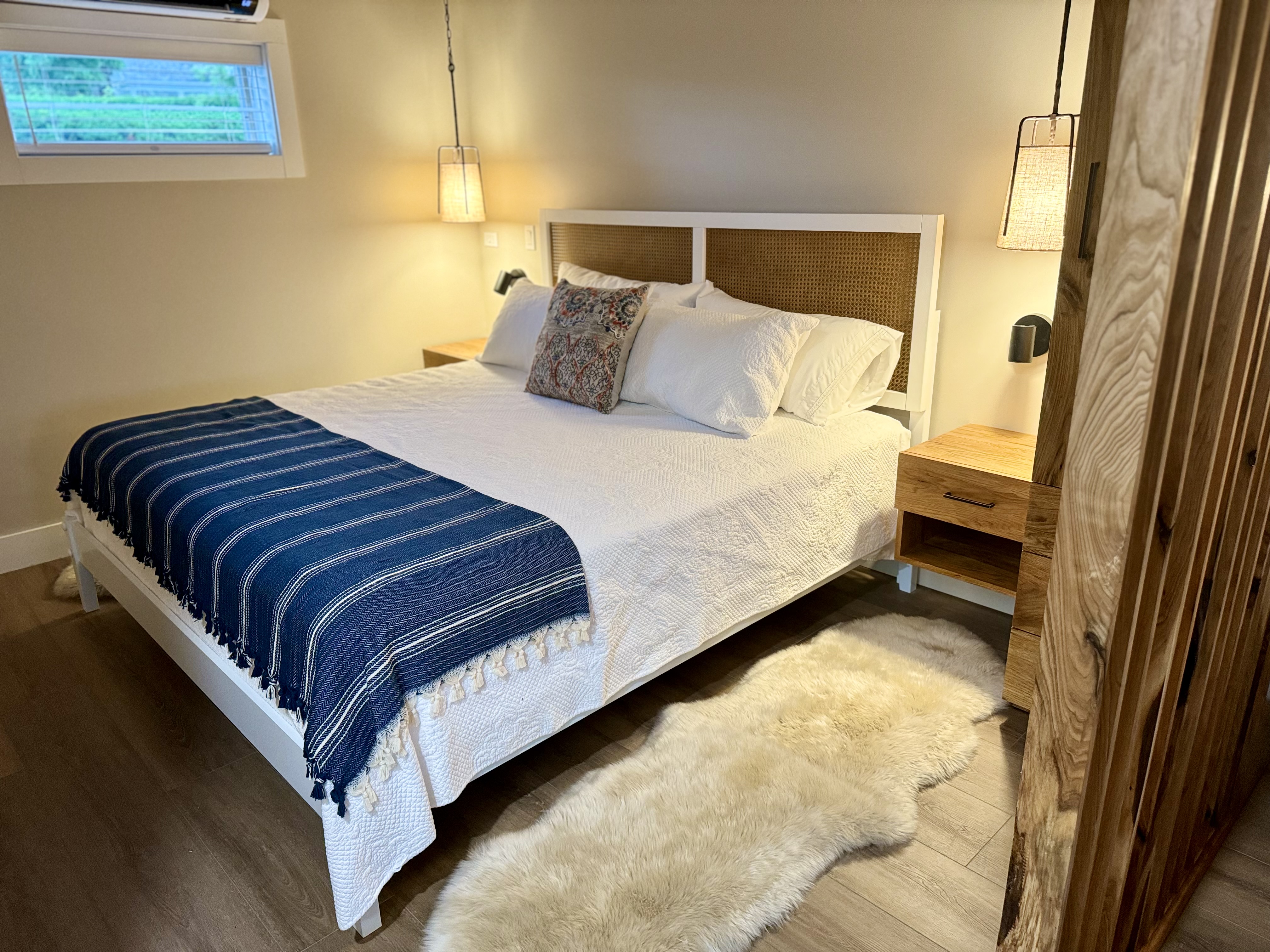 A bright and inviting bedroom features a king bed with a blue striped throw blanket, a woven headboard, and matching wood nightstands.