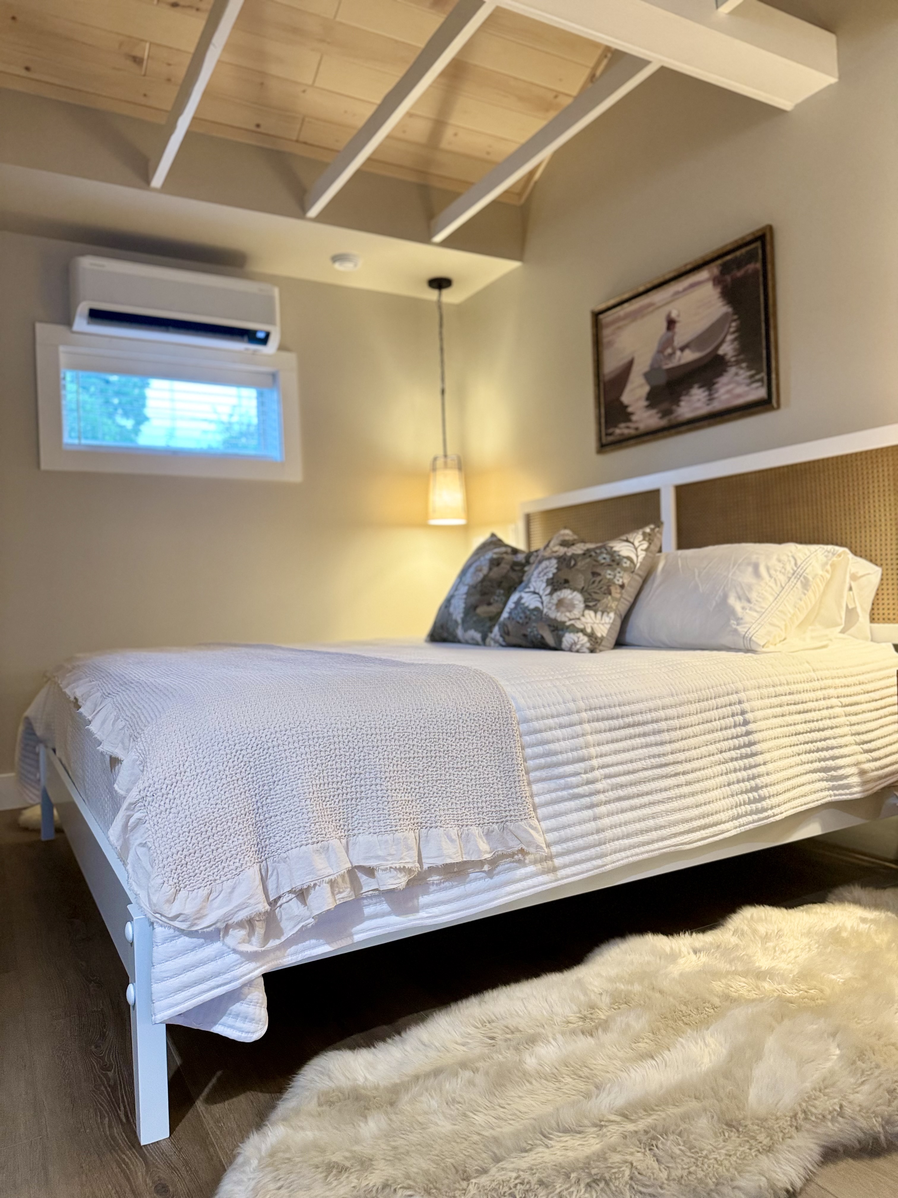 A bright and cozy bedroom features a king bed with white linens, a woven headboard, wood exposed beams on the ceiling, and a furry rug.