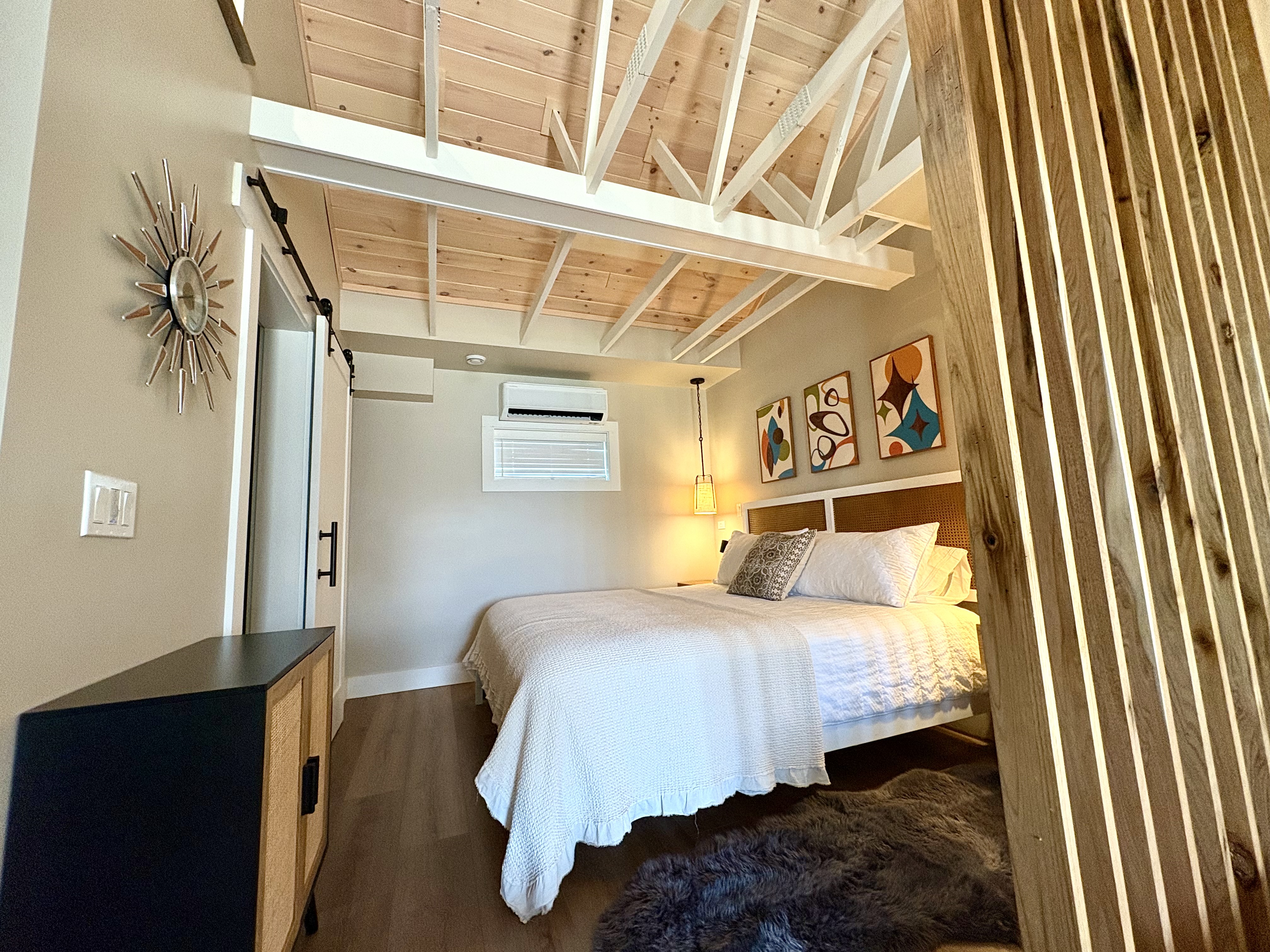 A light and inviting bedroom features a bed with white linens, a vaulted wood ceiling with white beams, a sliding barn door, and a wood slat partition.