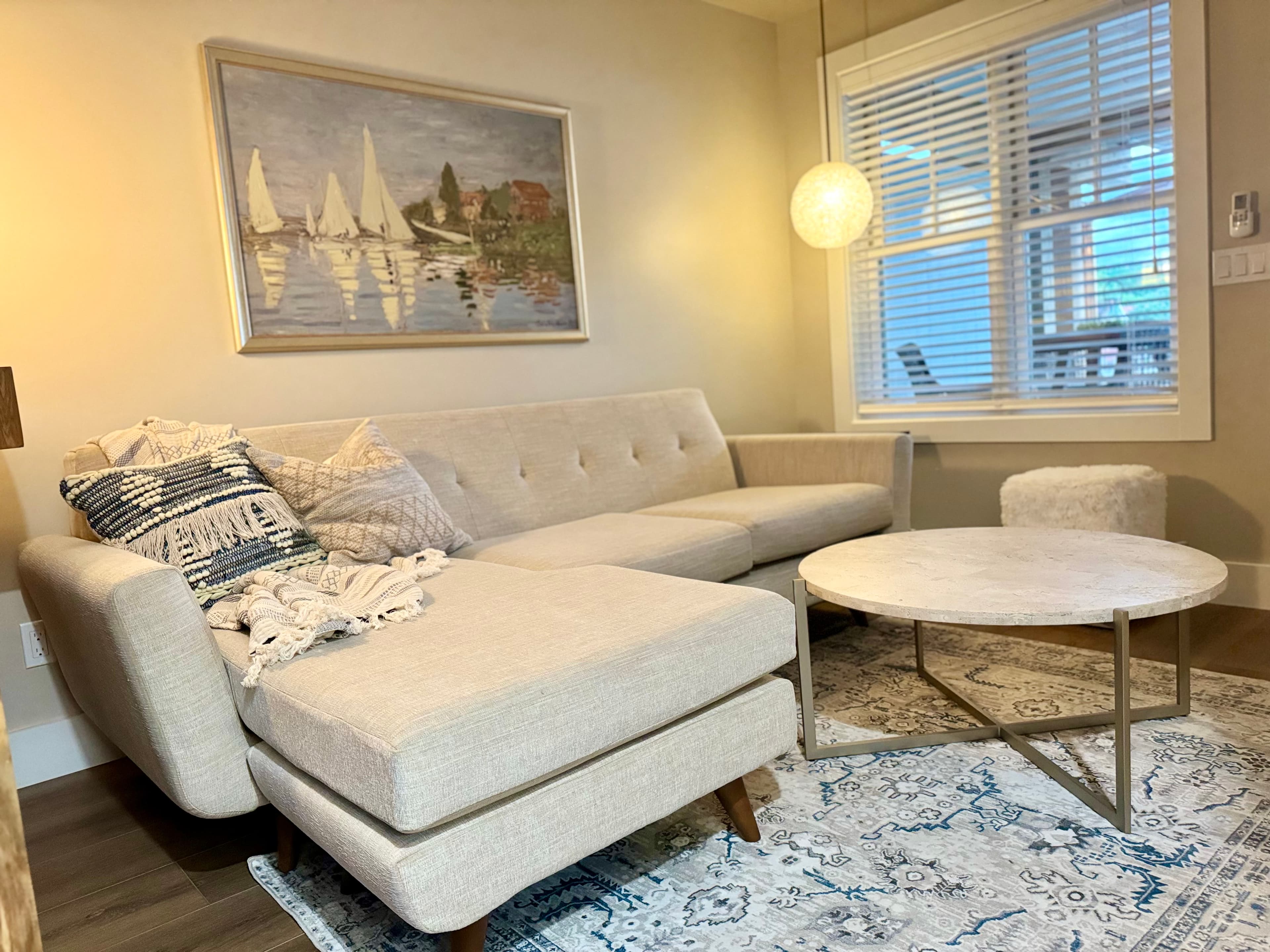 A bright living area featuring a beige sectional couch with patterned throw pillows, a round marble coffee table, and sailboat artwork.