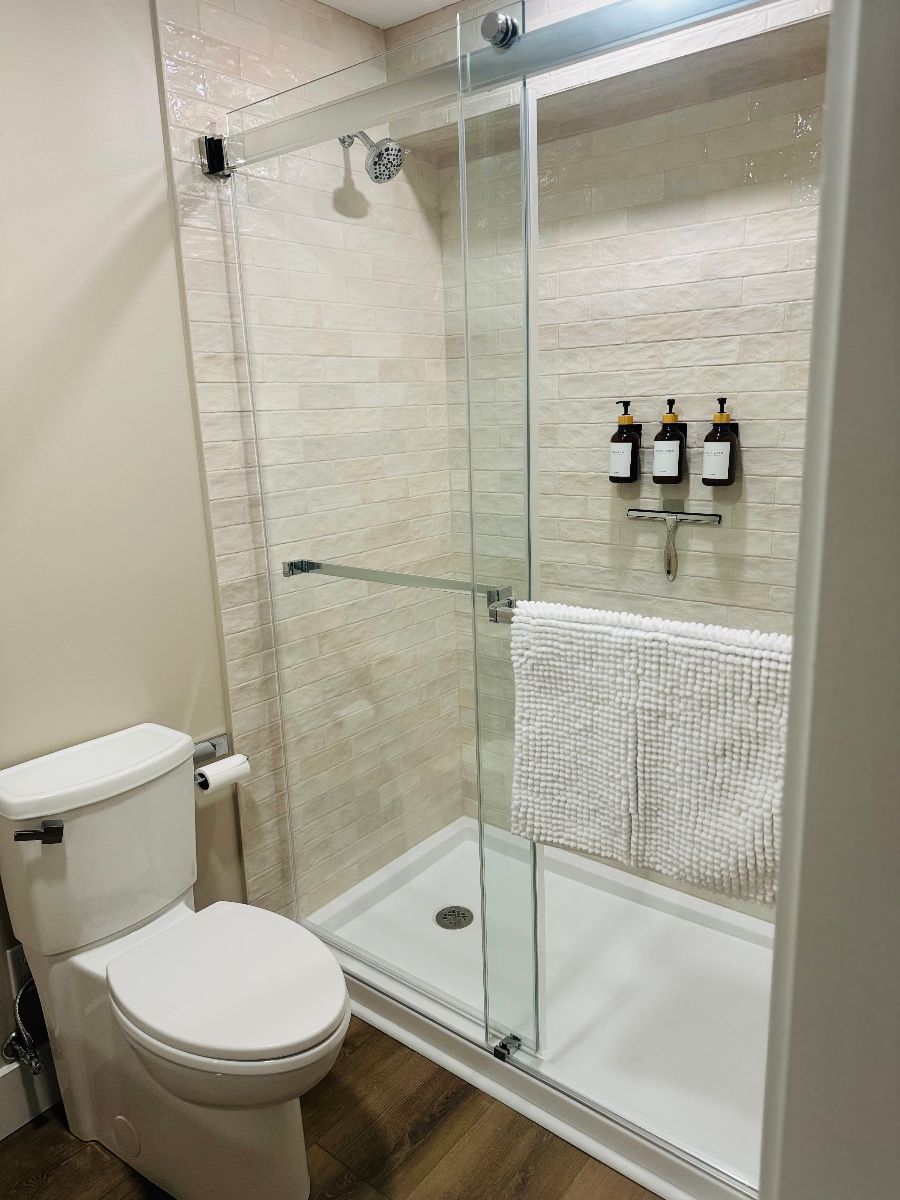 A modern bathroom featuring a walk-in shower with a glass door, light textured tile, and a white toilet.