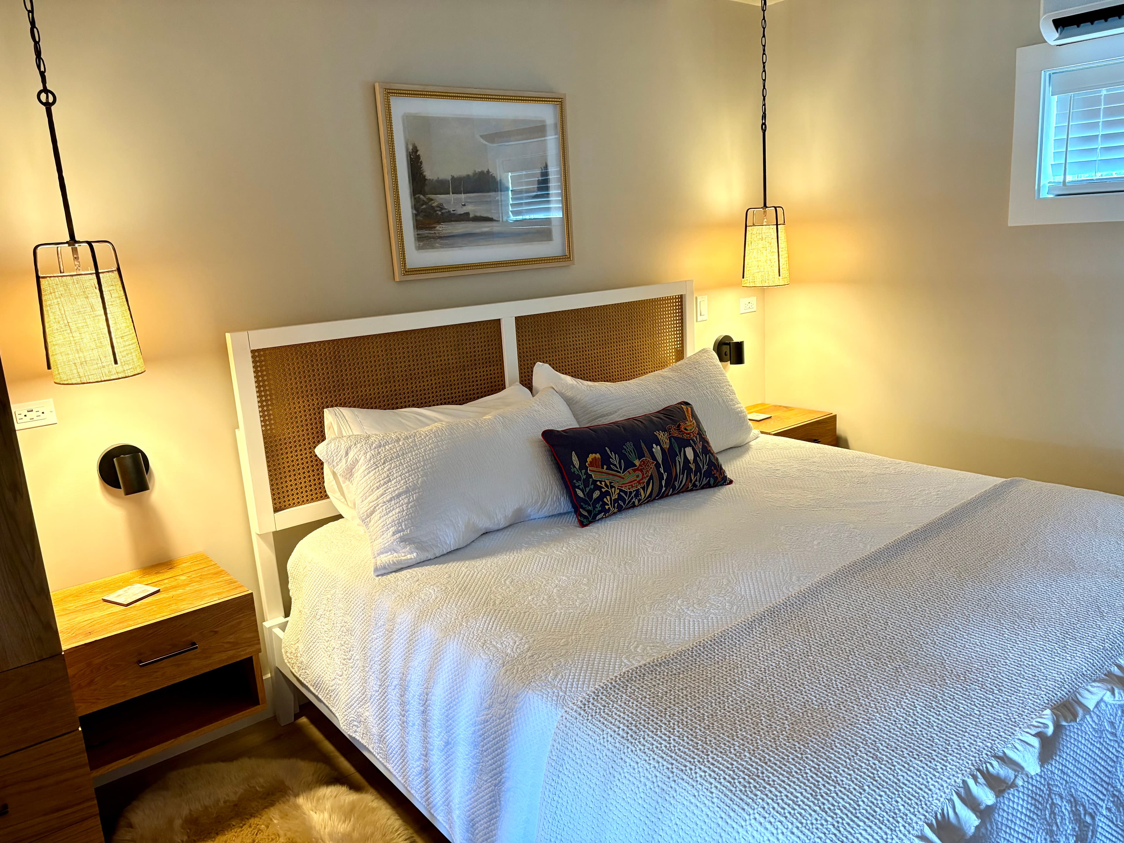 A warm and cozy bedroom features a king bed with white linens, a woven headboard, matching wood nightstands, and stylish pendant lights.