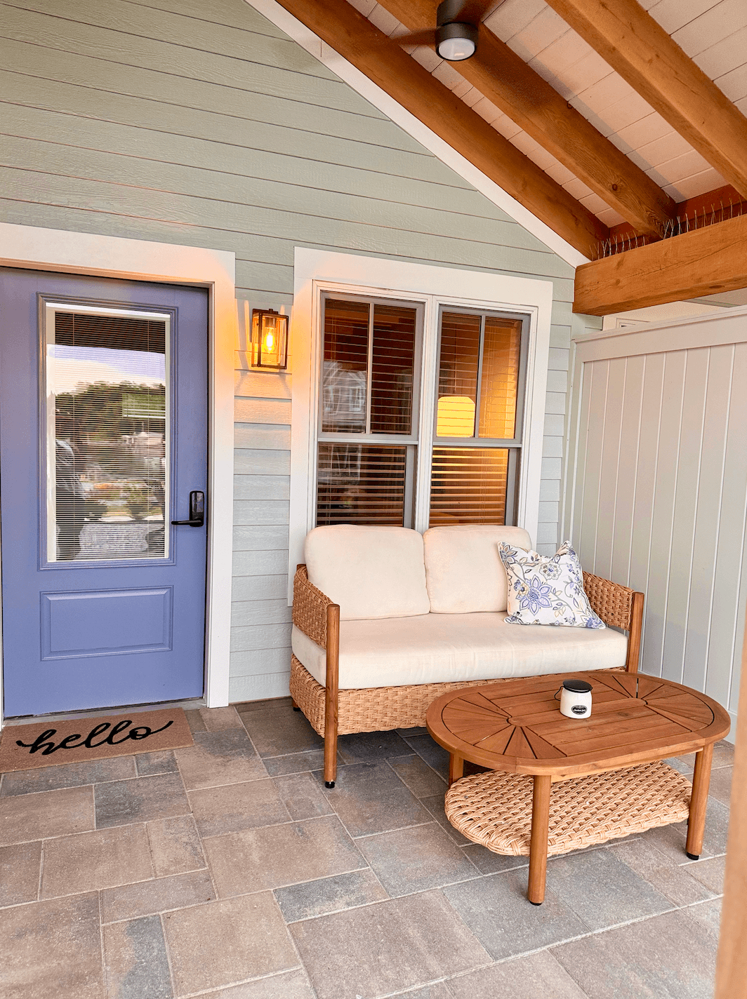 A welcoming covered porch features a light blue door, a wicker loveseat with white cushions, and a wood coffee table on a tile floor.