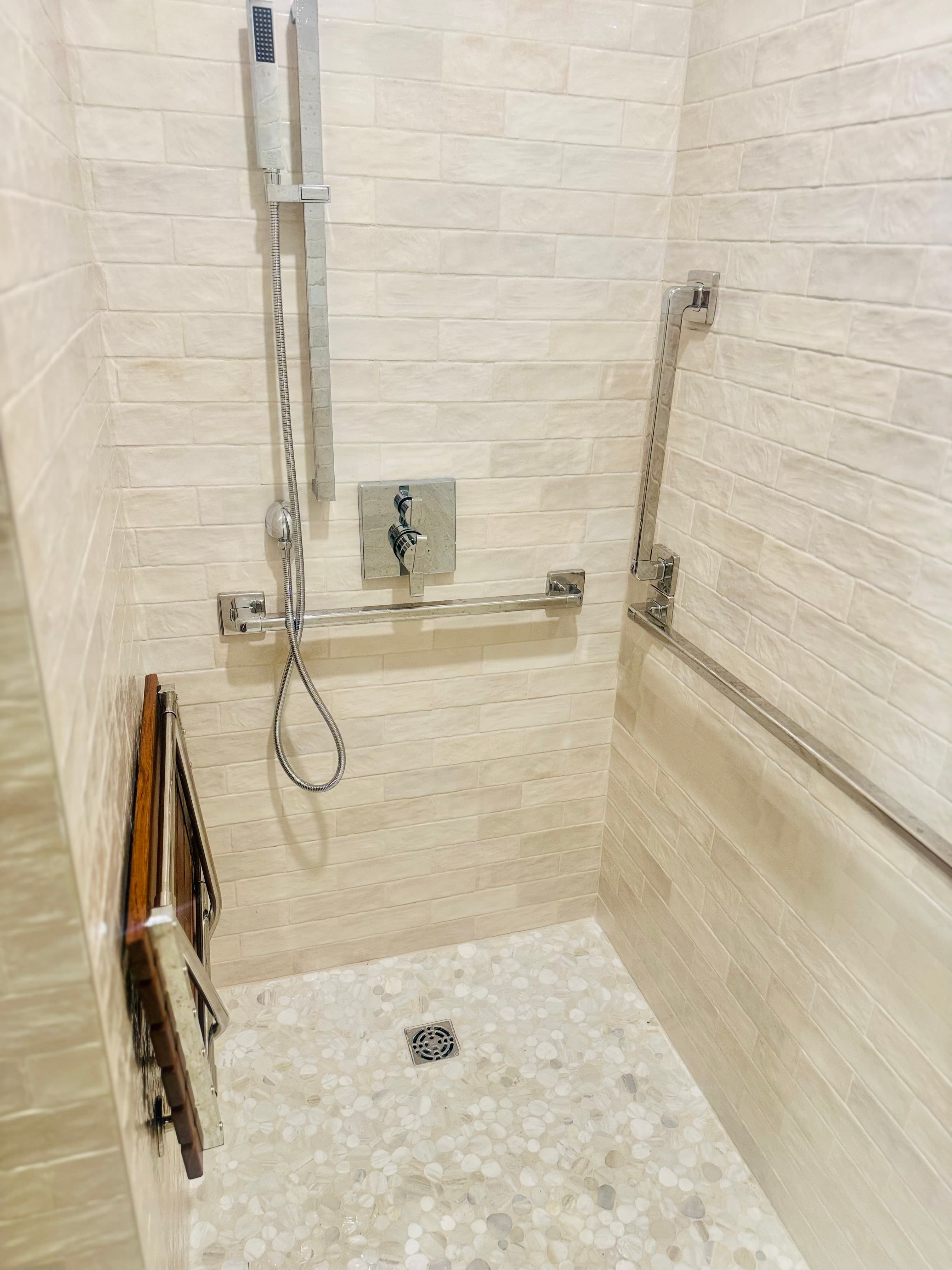 A modern, accessible walk-in shower features light-colored tile walls, a pebble floor, and a fold-down seat.