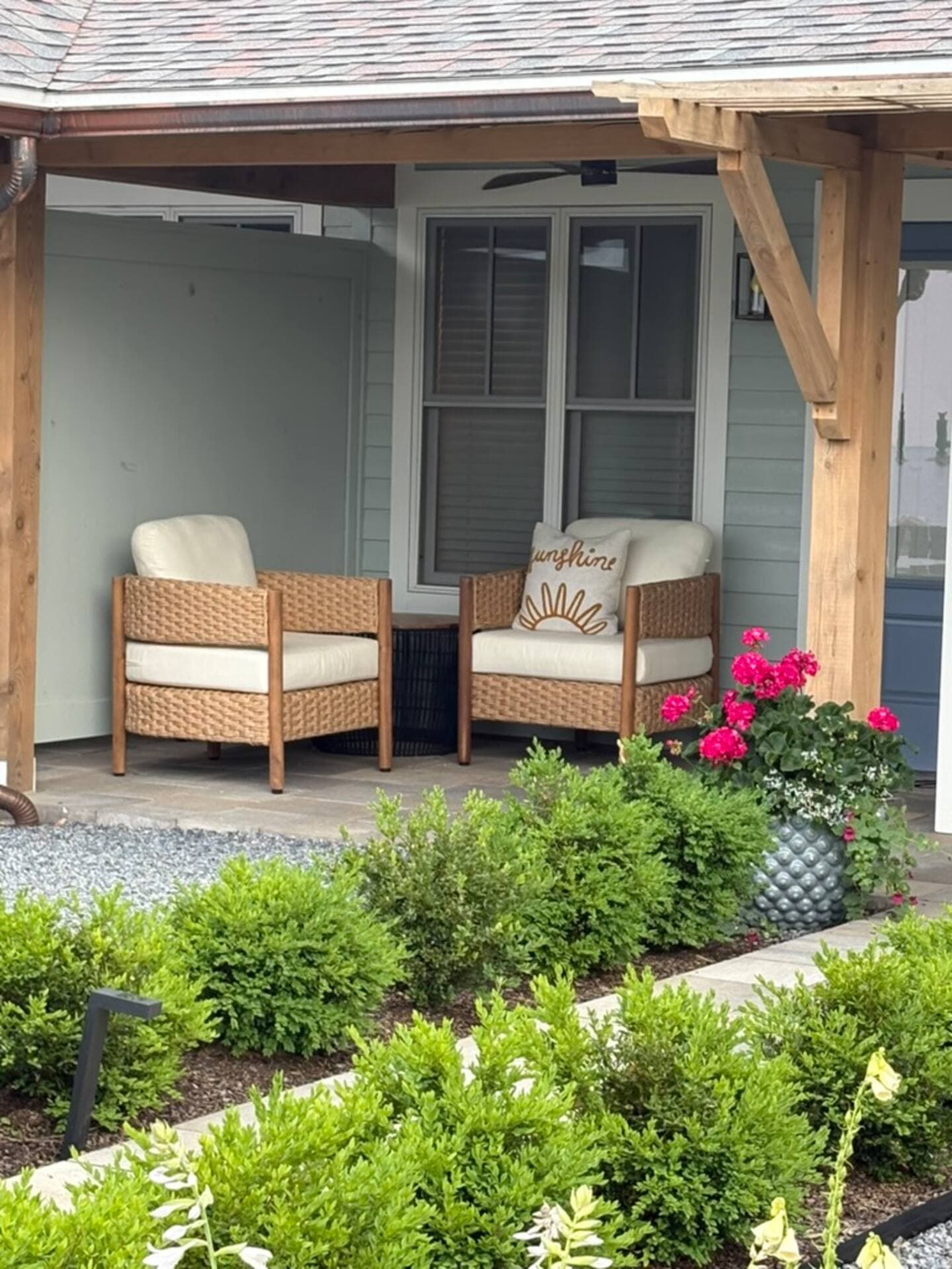 A serene covered patio features a pair of modern wood and cushion armchairs, colorful pink flowers, and a lush green garden in the foreground.