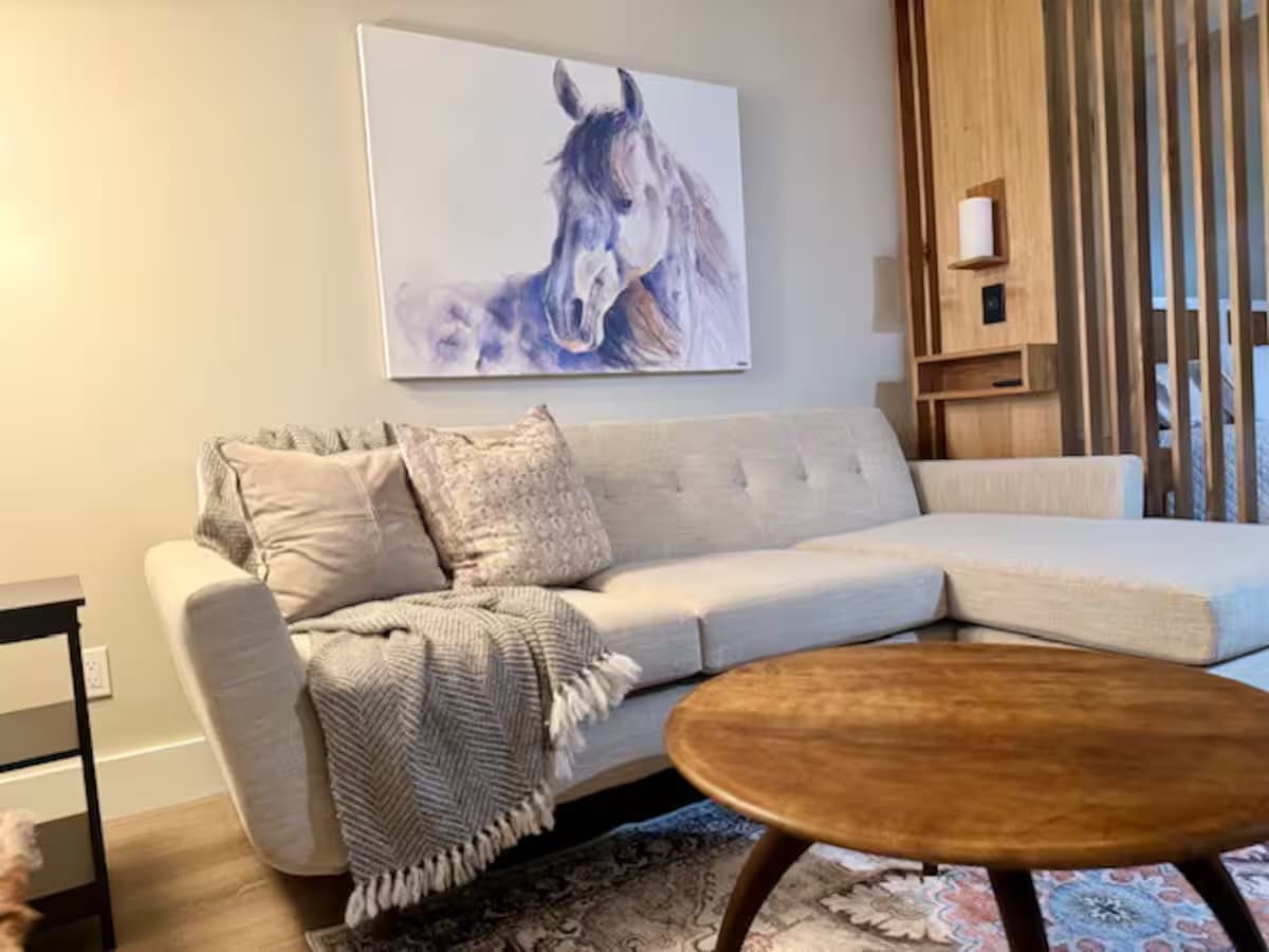 A cozy living area with a beige sectional sofa, a round wood coffee table, and an equestrian painting hanging above the couch.