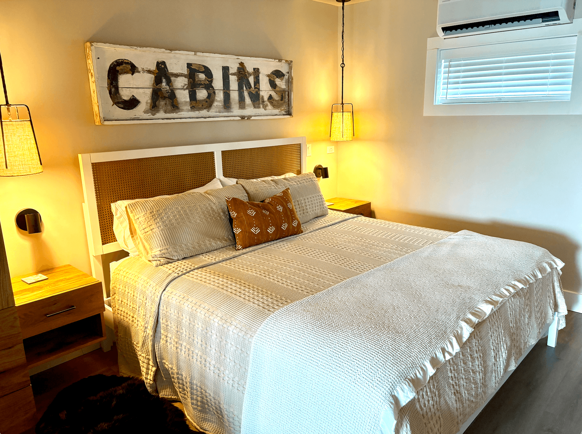 A cozy cottage bedroom features a bed with neutral linens, a woven headboard, and a rustic "CABINS" sign above.