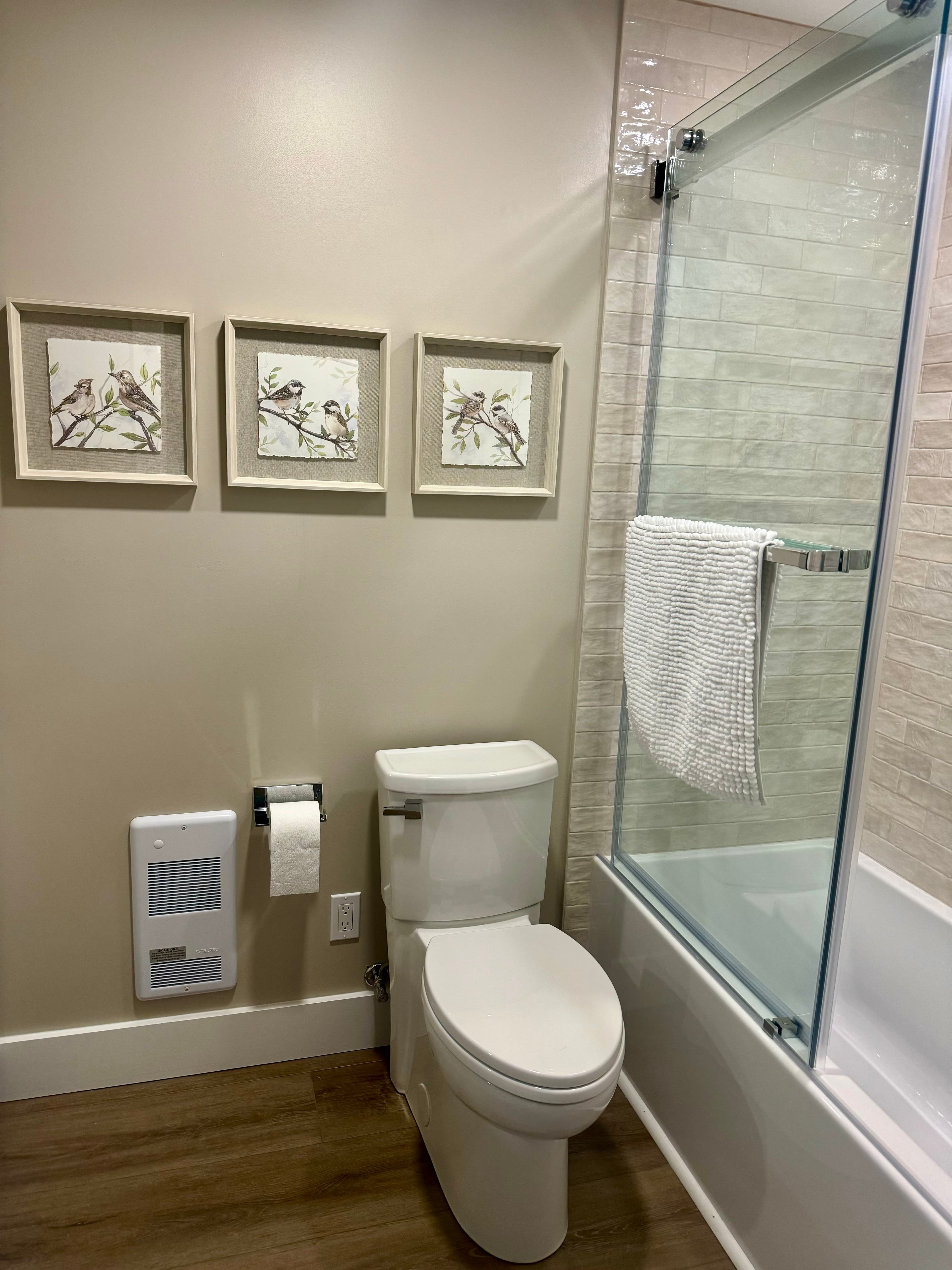 A modern bathroom with a white toilet next to a shower/soaking tub with a glass door. Three framed bird prints decorate the wall.