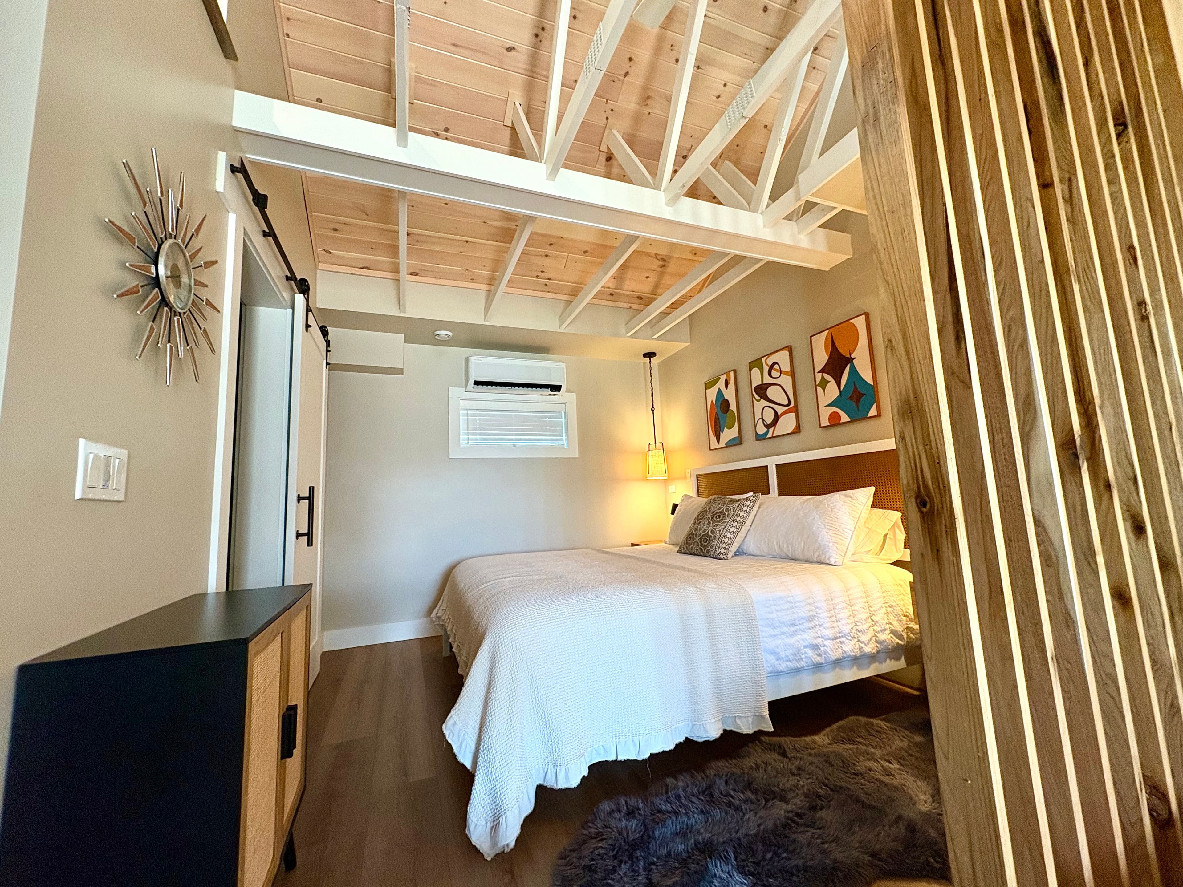 A light and inviting bedroom features a bed with white linens, a vaulted wood ceiling with white beams, a sliding barn door, and a wood slat partition.