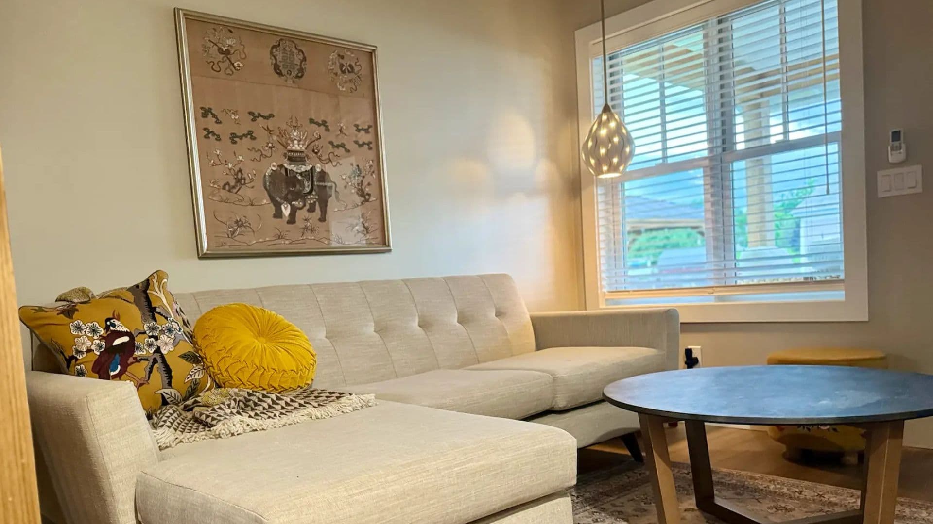 A cozy living room featuring a light gray sectional sofa, colorful throw pillows, a round coffee table, and a decorative wall hanging.
