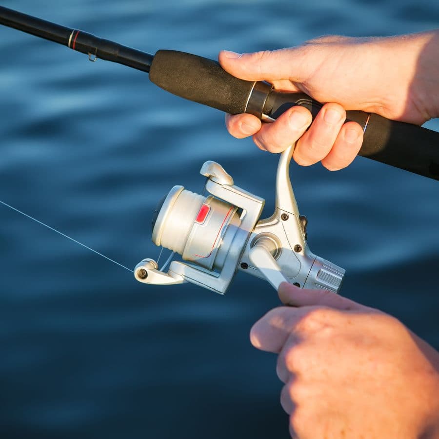 A person holds a fishing rod and reel with fishing line against a blurred water background.