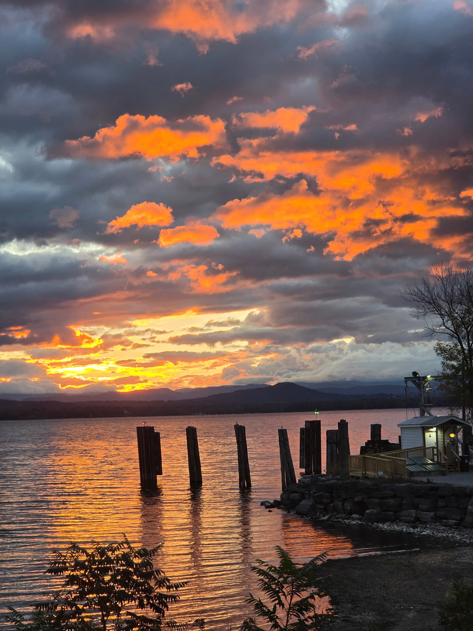 A vibrant sunset over a calm lake, with silhouetted wooden posts and dark clouds.