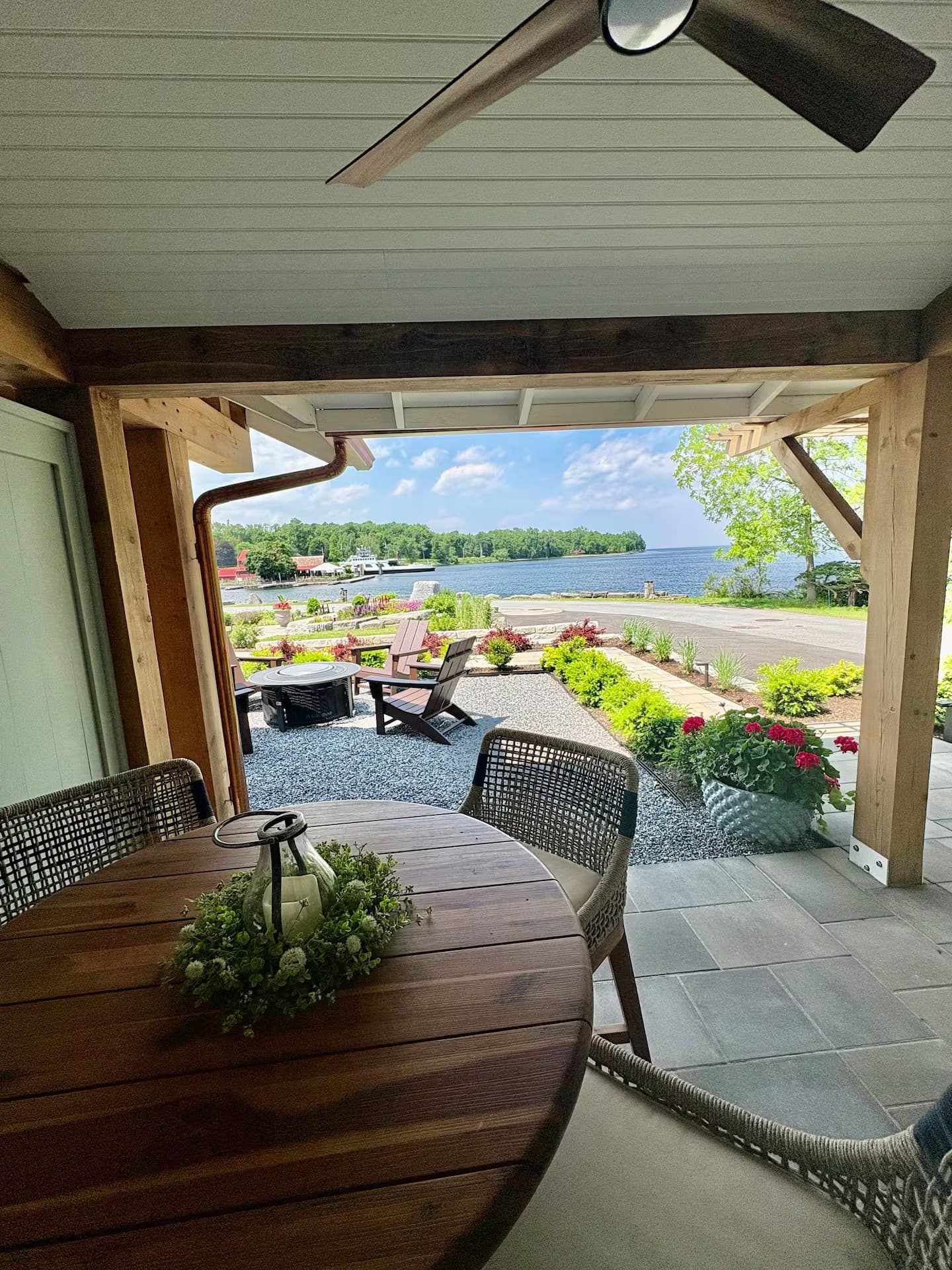 View of a lakeside patio with a round wooden table, chairs, and colorful garden flowers.