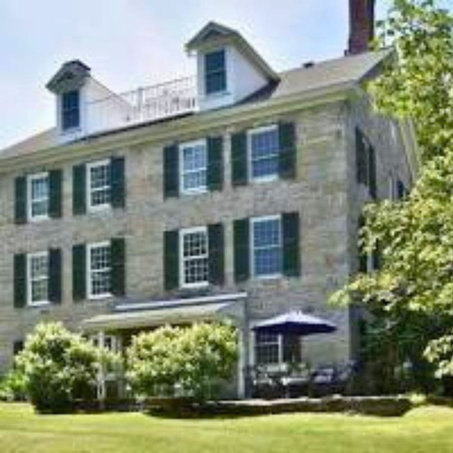 A historic stone house with green shutters and a patio surrounded by greenery.