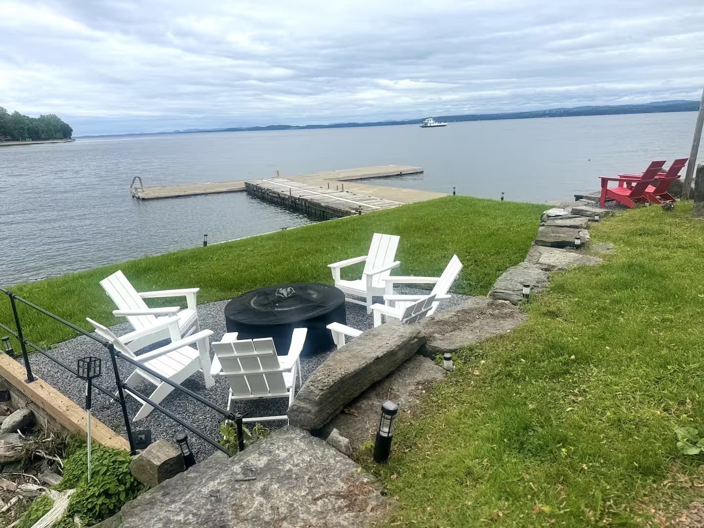 Lawn chairs surround a fire pit near a dock on a calm lake under a cloudy sky.