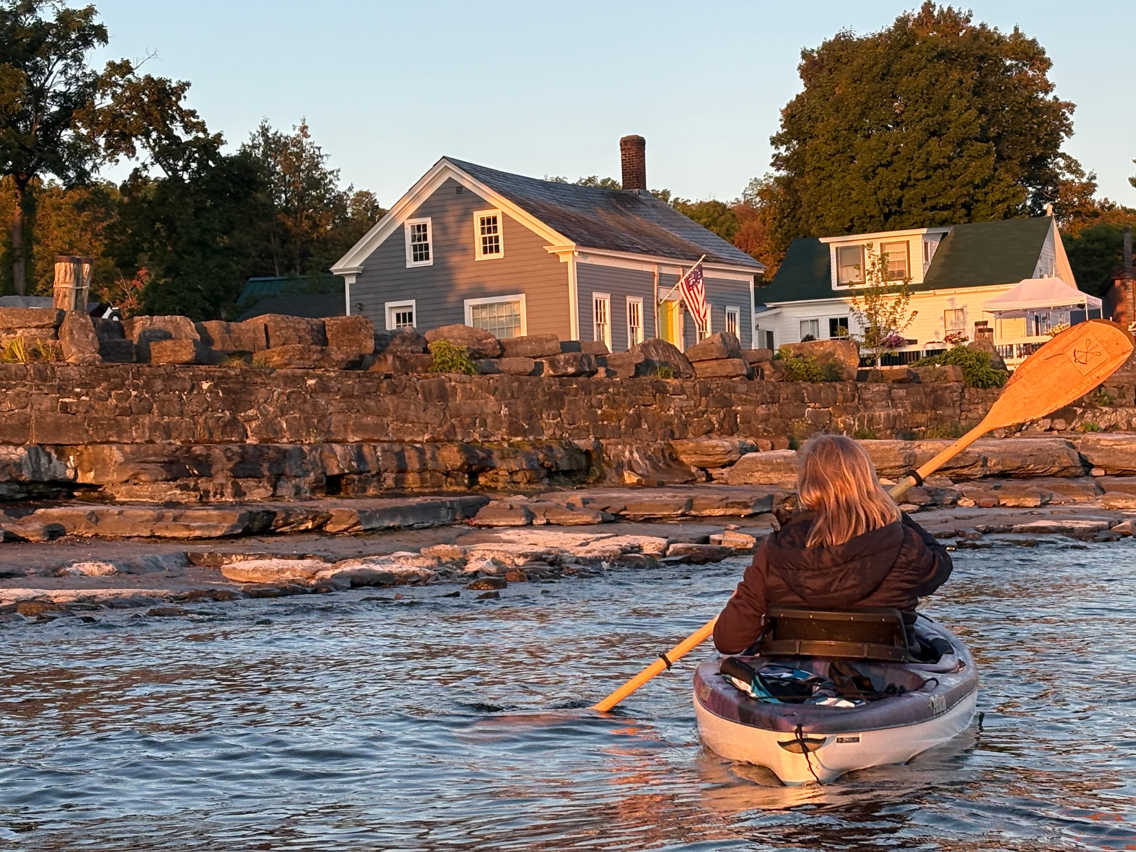 A person paddles a kayak past a shoreline house at sunset.