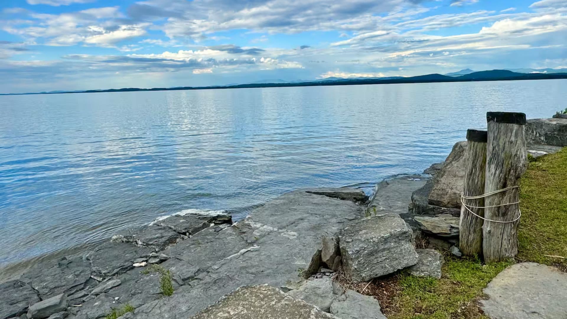 A serene lakeside view with smooth rocks, gentle waves, and a cloudy blue sky.