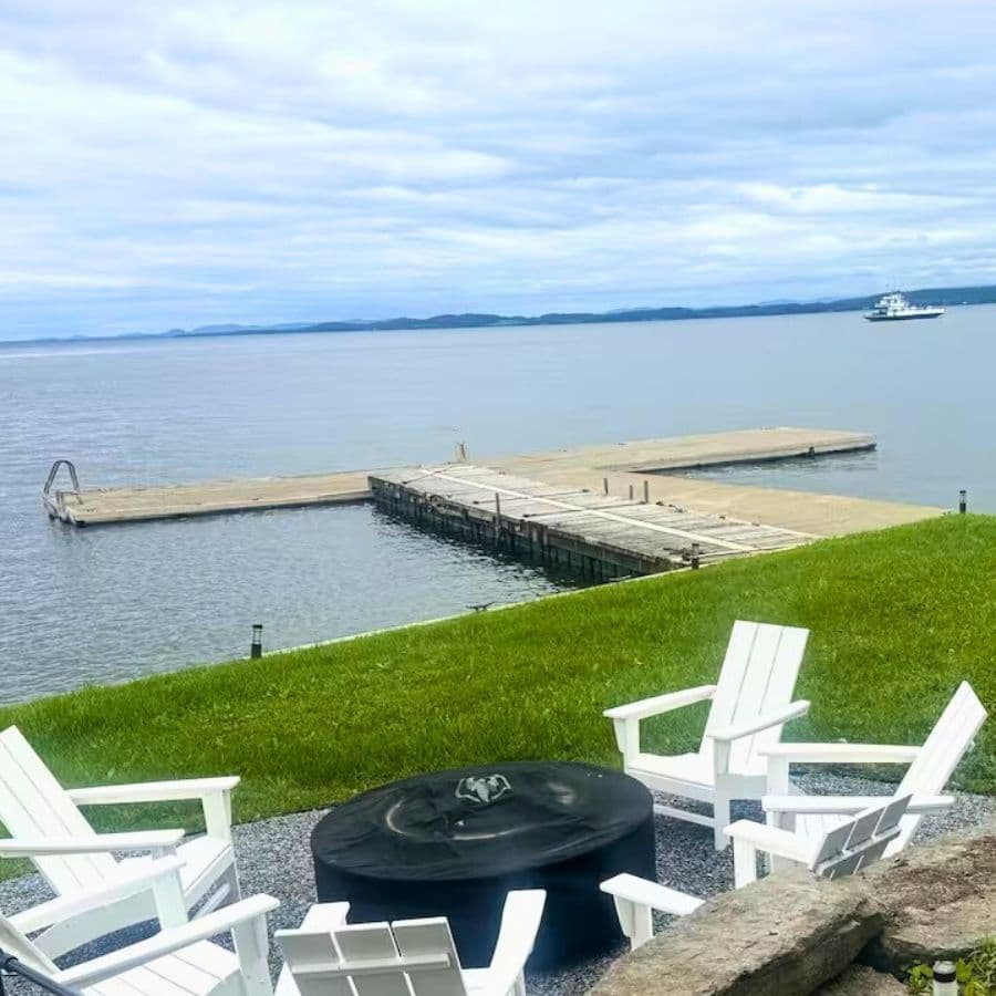 A tranquil lakeside view featuring a dock, green grass, and white chairs around a fire pit.