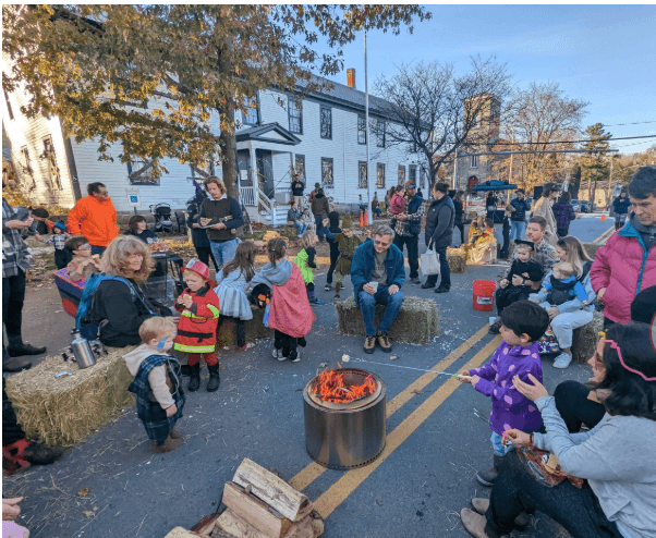 A crowd gathers around a central fire pit during a community event, with families and children engaging in activities outside a historic building. A crowd gathers around a central fire pit during a community event, with families and children engaging in activities outside a historic building.