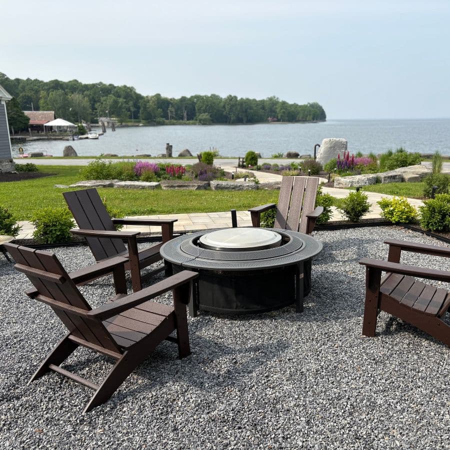 A circular fire pit surrounded by wooden chairs, overlooking a serene lake and lush greenery.