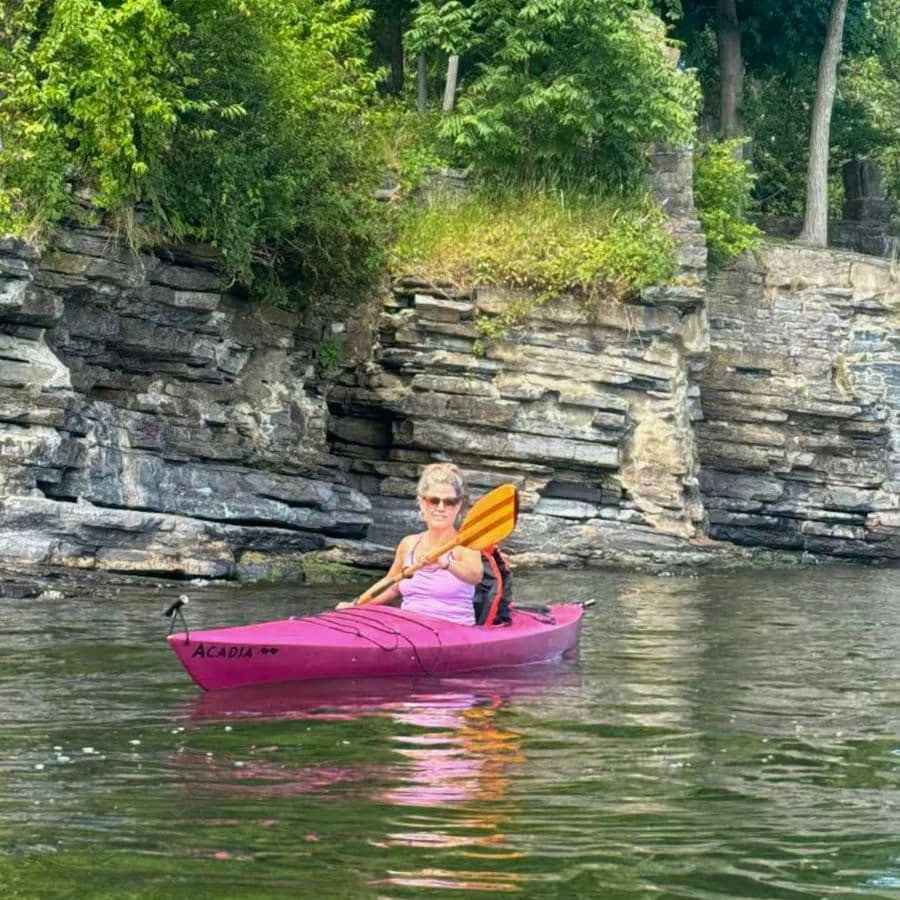 A woman in a pink kayak paddles on a calm body of water near rocky shorelines.