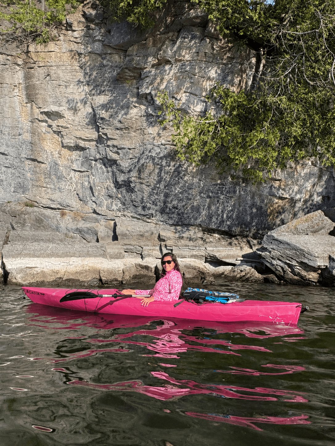 A person in a pink kayak sits on calm water beside a rocky shoreline.