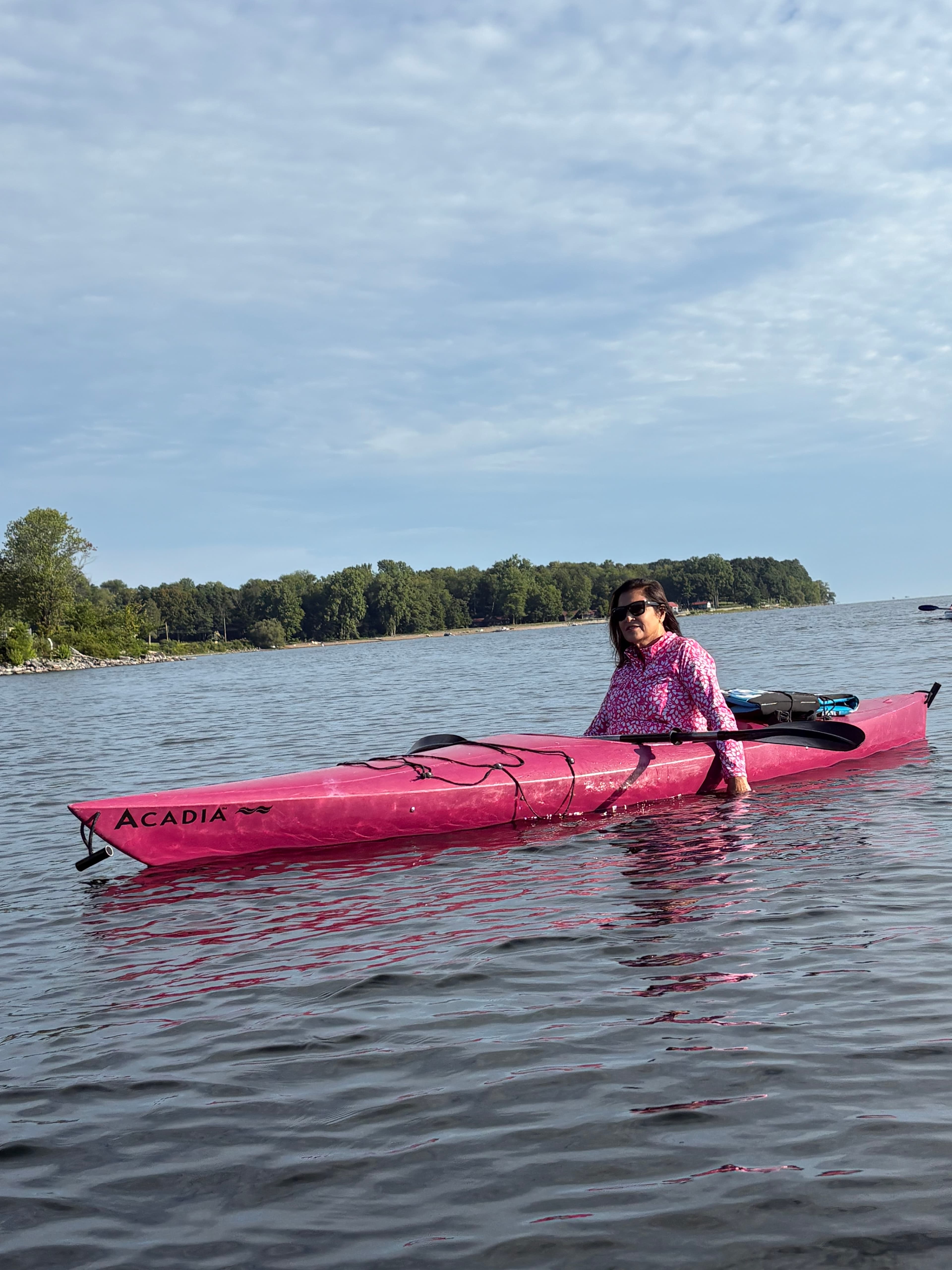 A person in a pink kayak is enjoying a calm day on a lake, surrounded by trees and a cloudy sky.