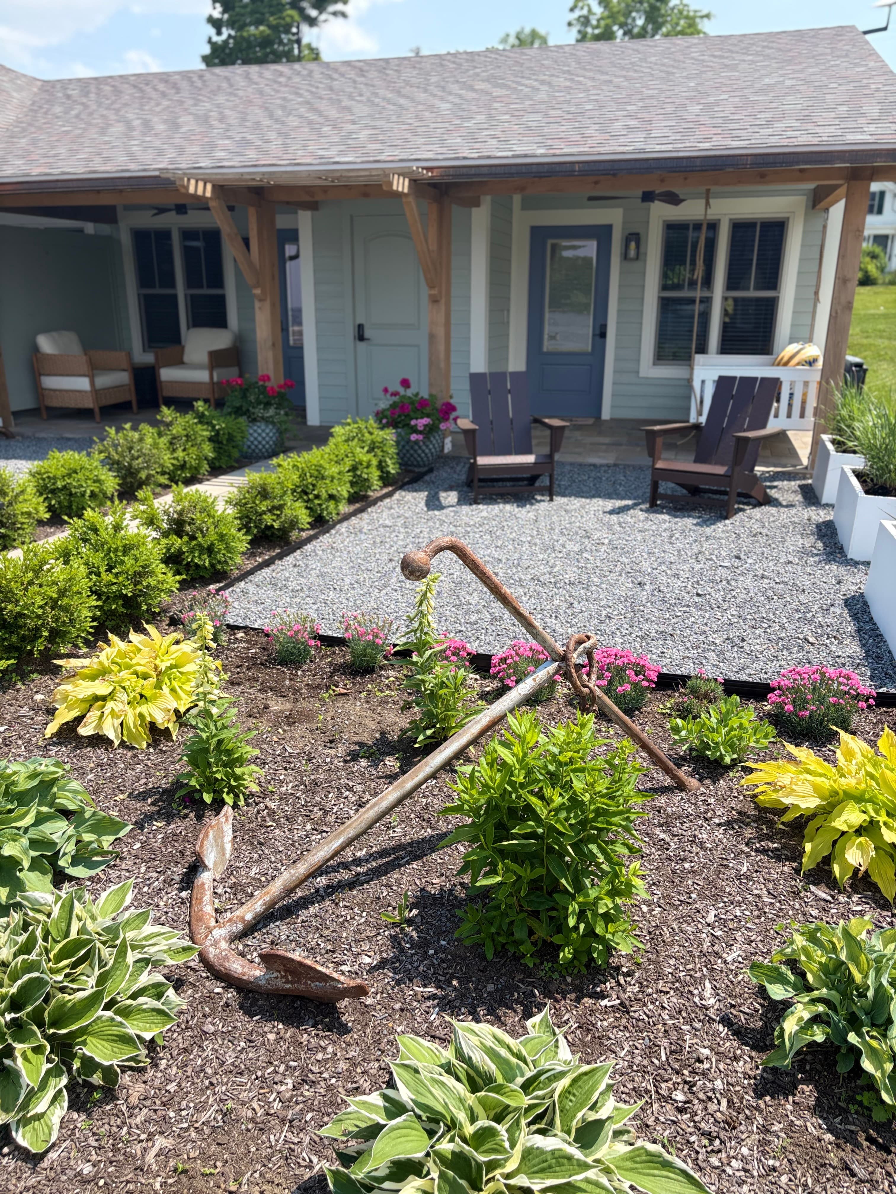 A rusted anchor is nestled among vibrant garden plants in front of a charming cottage.